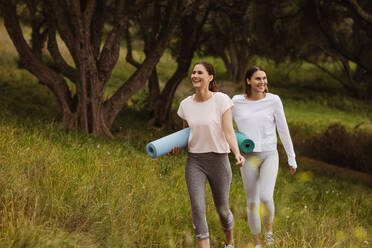 Zwei fröhliche Frauen, die mit Yogamatten in einem Park spazieren gehen. Lächelnde Freundinnen, die im Freien trainieren. - JLPSF12400