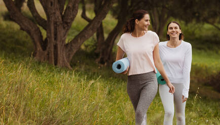 Fröhliche Fitness-Frauen genießen ihren Spaziergang in einem Park mit Yogamatten. Frauen, die morgens im Freien trainieren. - JLPSF12403