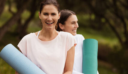 Zwei lächelnde Fitness-Frauen stehen im Park und halten Yogamatten in der Hand. Frauen gehen zum Training ins Freie. - JLPSF12405