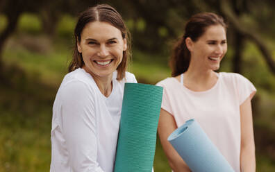 Fröhliche Freundinnen stehen im Freien und halten Yogamatten. Glückliche Fitness-Frauen beim Training. - JLPSF12406