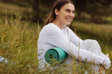 Fröhliche Frau mit Fitnessmatte im Park sitzend. Nahaufnahme einer lächelnden Frau, die sich nach einem Workout auf einer Wiese entspannt. - JLPSF12414
