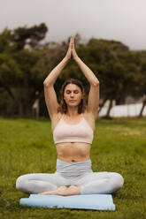 Fitness-Frau auf dem Boden sitzend mit verbundenen Handflächen über dem Kopf in einer Yoga-Pose. Porträt einer Frau, die im Park sitzt und Yoga praktiziert. - JLPSF12420