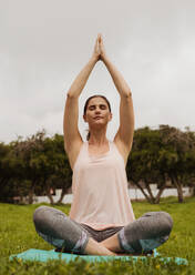 Close up of a fitness woman sitting in a yoga pose in a park. Woman practising yoga sitting in a park with palms joined together over her head. - JLPSF12429