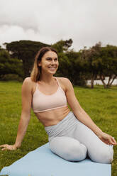 Smiling fitness woman sitting in a park looking away. Portrait of a cheerful woman sitting in grass enjoying nature after workout. - JLPSF12430