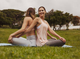 Fröhliche Fitness-Frauen beim Workout in einem Park. Freundinnen sitzen auf einer Yogamatte und machen Dehnübungen in entgegengesetzter Richtung. - JLPSF12434