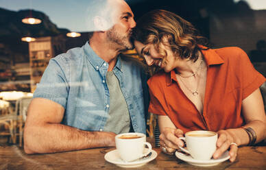 Mann küsst die Stirn seiner Frau, die im Café sitzt. Verliebtes Paar bei einem Kaffee-Date. - JLPSF12451