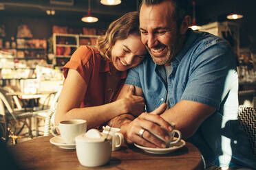Frau lächelt und legt ihren Kopf auf die Schulter eines Mannes, während sie in einem Café sitzt. Verliebtes Paar bei einem gemeinsamen Kaffee-Date. - JLPSF12453