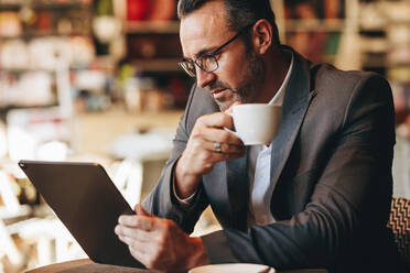 Geschäftsmann, der mit einer Tasse Kaffee in der Hand in einem Café auf sein digitales Tablet schaut. Geschäftsmann, der in einem Café an einem digitalen Tablet arbeitet - JLPSF12460