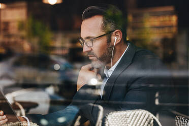 Älterer Geschäftsmann in einem Café mit Kopfhörern, der auf sein digitales Tablet schaut. Geschäftsmann, der einen Videoanruf von seinem Tablet-Computer macht, während er in einem Café sitzt. - JLPSF12461