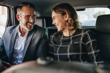 Two business people traveling in backseat of a car. Businessman and businesswoman looking at each other and smiling while riding a taxi to work. - JLPSF12466