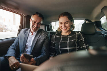 Businesswoman working on laptop with a businessman using mobile phone while traveling by a car. Business people working in backseat of taxi. - JLPSF12467