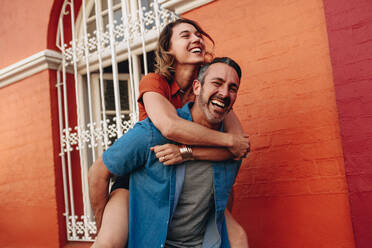 Mature man carrying his girlfriend on his back walking on the city street. Couple piggybacking on city street. - JLPSF12477