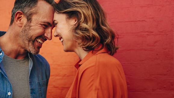 Beautiful couple in love touching foreheads and smiling. Happy couple standing against red and orange colored wall. - JLPSF12479