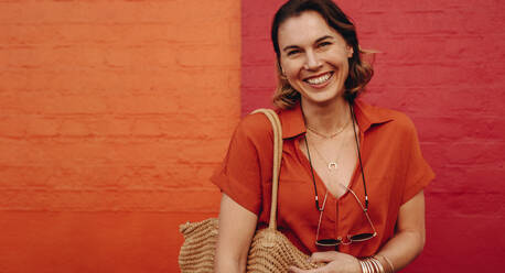 Porträt einer schönen Frau, die vor einer roten und orangefarbenen Wand steht, in Freizeitkleidung, die in die Kamera schaut und lächelt. - JLPSF12483
