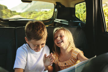 Geschwister sitzen auf der Rückbank eines Autos und schauen sich lächelnd eine Landkarte an. Kinder, die in einem Auto auf einem Roadtrip unterwegs sind und mit einer Karte spielen. - JLPSF12493