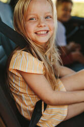 Close-up of a cute girl sitting in backseat of car wearing seatbelt. Cheerful girl enjoying travelling by a car. - JLPSF12497