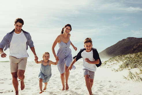 Glückliche junge vierköpfige Familie, die gemeinsam am Strand läuft. Eltern mit Kindern, die am Strand entlang laufen. - JLPSF12501