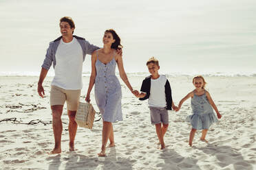 Family of four taking a walk along the sea shore with a picnic basket. Couple with their children walking on beach. - JLPSF12511