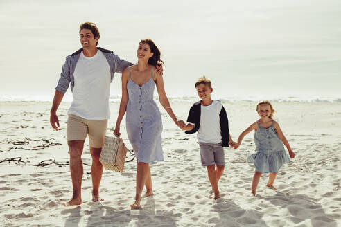 Vierköpfige Familie, die mit einem Picknickkorb am Meer spazieren geht. Ehepaar mit ihren Kindern, die am Strand spazieren gehen. - JLPSF12511
