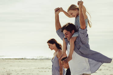 Happy family on a summer vacation at beach. Man giving daughter ride on shoulders at the seashore while walking with family.. - JLPSF12515