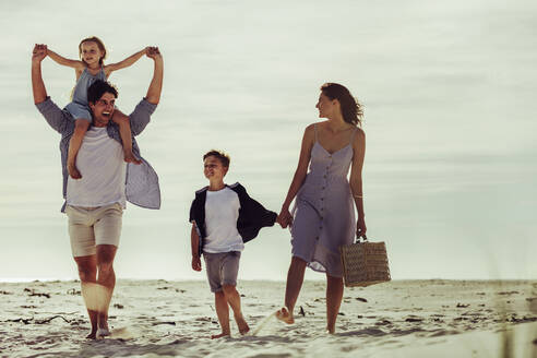 Vierköpfige Familie, die mit einem Picknickkorb am Meer entlang spaziert. Ehepaar mit ihren beiden Kindern, die am Strand spazieren gehen. - JLPSF12516