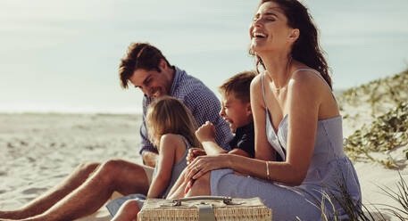 Familie sitzt am Strand und lächelt. Frau mit ihrer Familie entspannt sich am Strand und lacht. - JLPSF12517