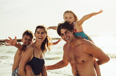 Glückliche Mutter und Vater, die ihre Kinder am Strand huckepack nehmen. Zwei Kinder, die mit ihren Eltern im Sommerurlaub Spaß haben. - JLPSF12526