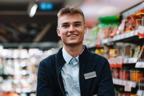 Porträt eines Mannes, der in einem Lebensmittelladen arbeitet und lächelnd in die Kamera schaut. Junger Arbeiter in Uniform im Supermarkt. - JLPSF12541