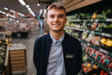 Nahaufnahme eines jungen männlichen Verkäufers. Ein Mann in Uniform, der im Supermarkt arbeitet, lächelt in die Kamera. - JLPSF12547
