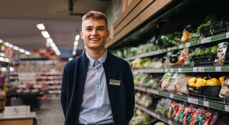 Portrait of a male worker at a grocery store and looking at the camera smiling. Man in uniform standing in supermarket. - JLPSF12549