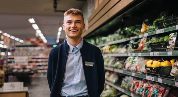 Porträt eines männlichen Angestellten in einem Lebensmittelgeschäft, der lächelnd in die Kamera blickt. Mann in Uniform im Supermarkt stehend. - JLPSF12549
