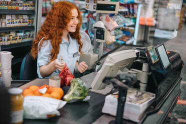 Young sales clerk sitting by cash register in supermarket and serving shoppers. Woman cashier scanning grocery products at checkout. - JLPSF12550