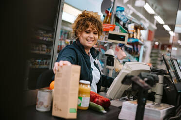 Freundliche Kassiererin, die die Produkte durch das Strichcodelesegerät im Supermarkt führt. Frau arbeitet an der Kasse eines Lebensmittelgeschäfts. - JLPSF12552