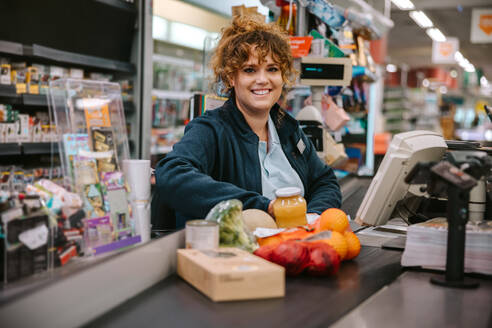 Portrait of a woman sitting behind checkout counter smiling at camera. Supermarket cashier at checkout. - JLPSF12553