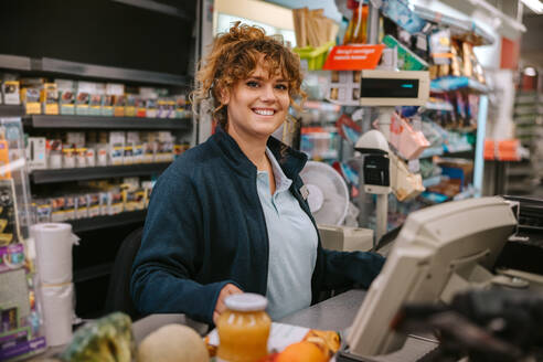 Porträt einer glücklichen Frau, die an der Kasse arbeitet und in die Kamera lächelt. Supermarktangestellte, die am Kassentresen arbeiten. - JLPSF12554