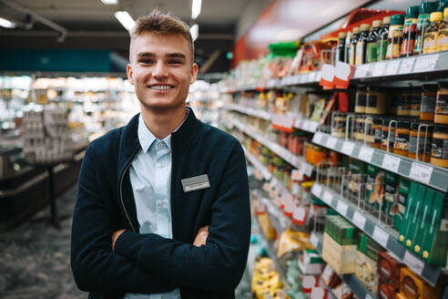 Porträt eines Mannes in Uniform, der mit verschränkten Armen in einem Lebensmittelgeschäft steht. Männlicher Arbeiter im Supermarkt. - JLPSF12562