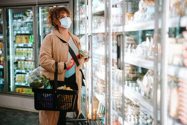 Frau mit Schutzmaske beim Einkaufen in einem modernen Supermarkt. Lebensmitteleinkauf während einer Pandemie. - JLPSF12566