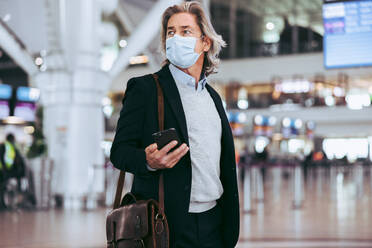 Mann mit Gesichtsmaske, Handtasche und Mobiltelefon auf einem Flughafen. Männlicher Geschäftsreisender im Transitbereich eines internationalen Flughafens. - JLPSF12589