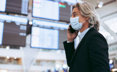 Man with protective face mask talking on cell phone while at airport. Mature businessman using phone at airport terminal. Air travel during covid-19 outbreak. - JLPSF12591