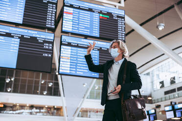 Man wearing face mask standing at airport terminal and waving at someone. Businessman traveling post pandemic lockdown. - JLPSF12592