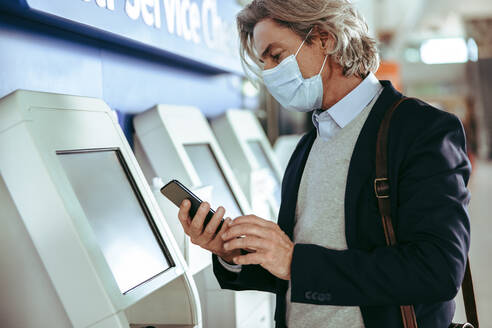 Männlicher Reisender mit Gesichtsmaske beim Self-Check-in an einem Automaten im Flughafenterminal und Überprüfung seines Mobiltelefons. Geschäftsmann beim Self-Check-in am Flughafen. - JLPSF12595