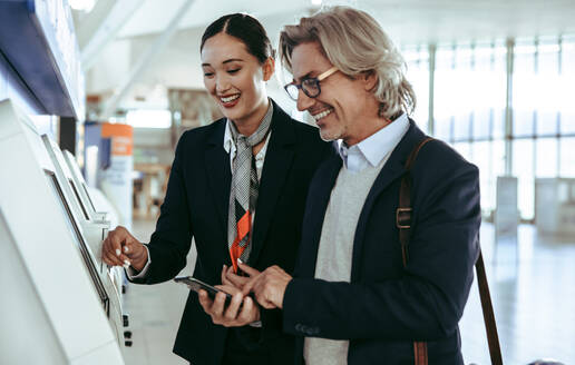 Freundliches Bodenpersonal hilft einem männlichen Reisenden beim Self-Check-in an einem Automaten im Flughafenterminal. Geschäftsmann am Self-Service-Check-in-Automaten mit weiblichem Flughafenpersonal. - JLPSF12599