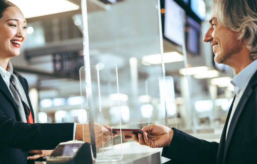 Geschäftsmann beim Einchecken im Flughafenterminal, der dem Bodenpersonal seinen Pass und sein Ticket übergibt. Transparente Plexiglaswand am Schalter zur Sicherheit. - JLPSF12603