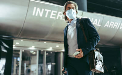 Businessman wearing face mask walking inside international airport. Business traveler with suitcase at airport during pandemic. - JLPSF12613
