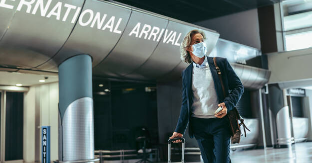 Mann auf Geschäftsreise geht mit seinem Gepäck am Flughafen spazieren. Reisender Geschäftsmann mit Gesichtsmaske. - JLPSF12615