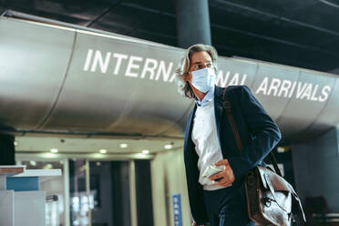 Businessman with face mask infront of international arrivals gate at airport. International business traveler at airport terminal post pandemic lockdown - JLPSF12616