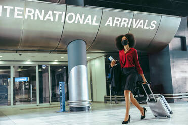 Businesswoman wearing face mask with luggage walking in airport terminal. International traveler arrived with suitcase during pandemic. - JLPSF12619