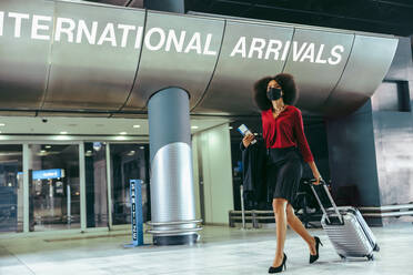 Geschäftsfrau mit Gesichtsmaske und Gepäck geht im Flughafenterminal spazieren. Internationaler Reisender kam mit Koffer während der Pandemie an. - JLPSF12619