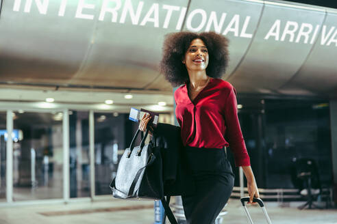 Lächelnde afrikanische Frau mit Gepäck auf dem internationalen Flughafen. Glückliche Frau auf internationaler Geschäftsreise. - JLPSF12620
