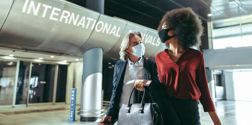 Two business traverlers wearing protective face masks walking together at airport terminal. Man and woman arriving at international airport during pandemic. - JLPSF12623